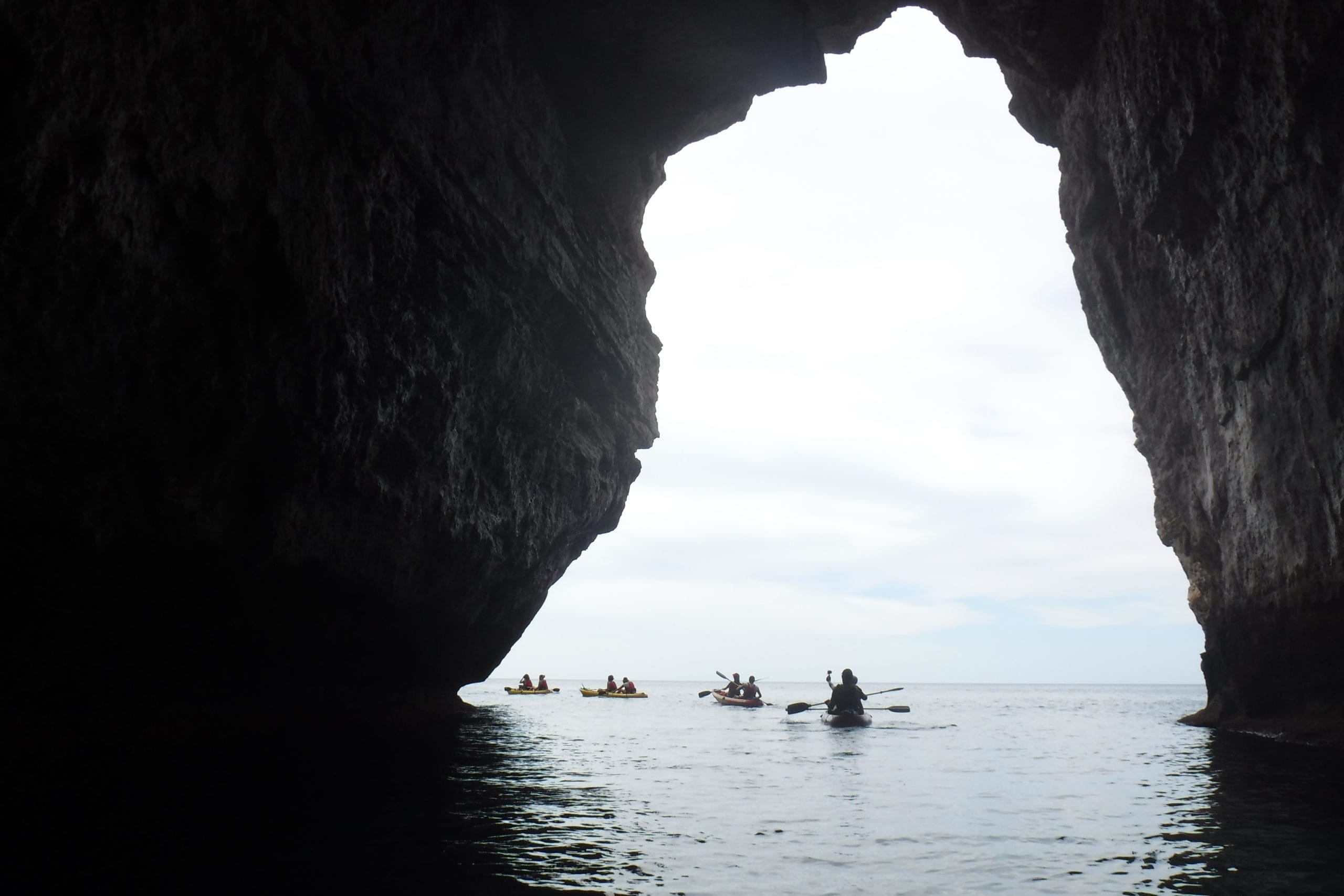 Conjunto de kayaks en la entrada de una cueva a contraluz. Recorrido por los acantilados de Cala En Porter y Cales Coves. Ruta guiada por Menorca en Kayak