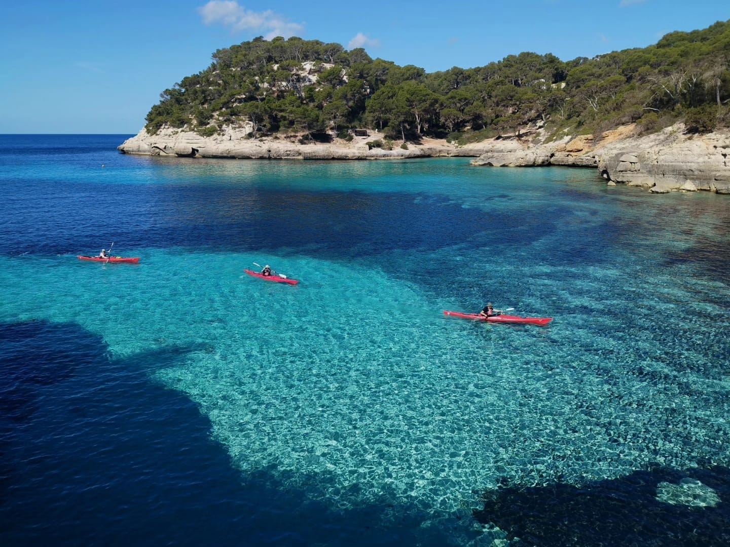 Tres kayaks de mar navegando por una cala paradisíaca de Menorca, rodeada de acantilados y bosque mediterráneo.