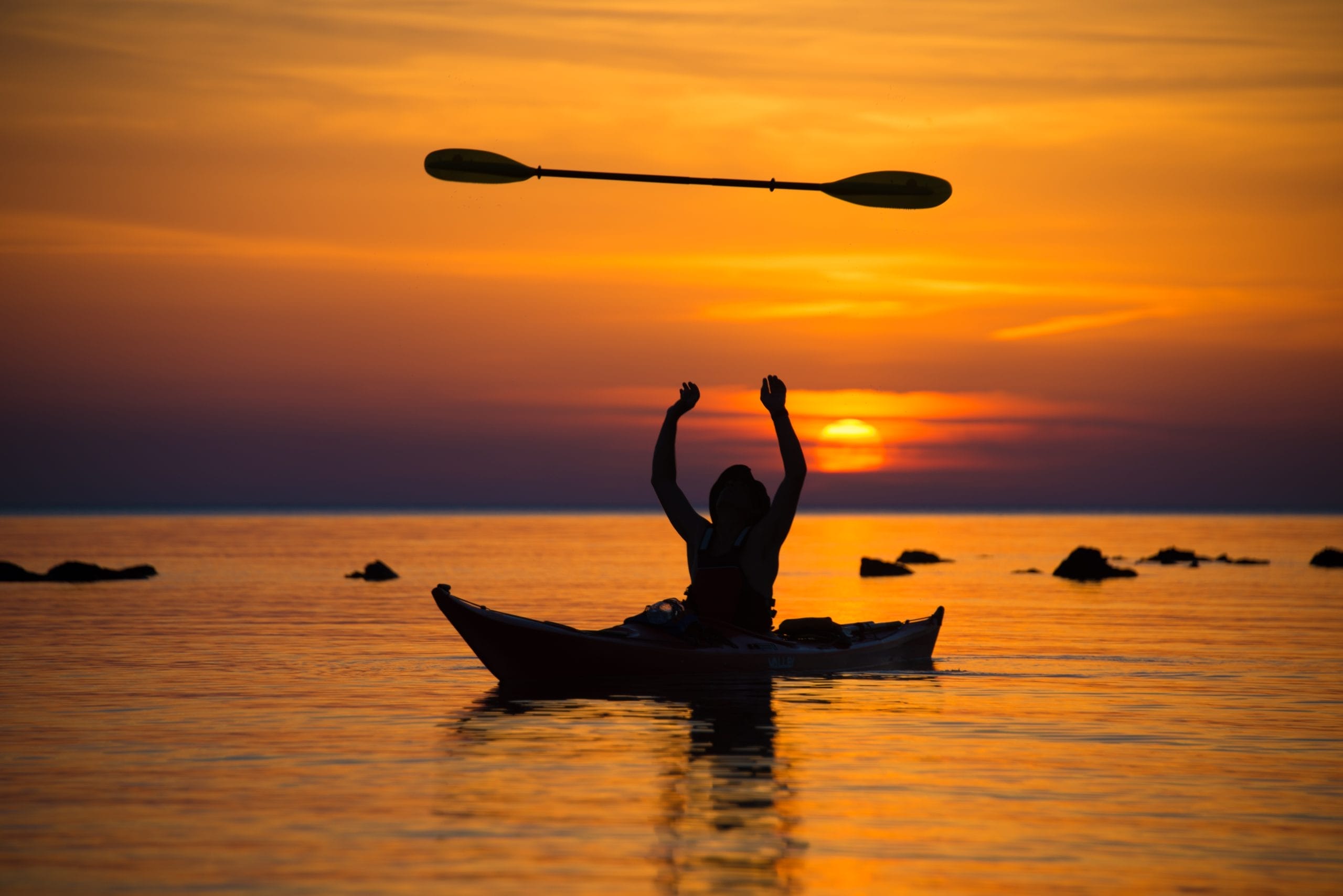 Silueta de una persona en kayak lanzando su pala al aire mientras disfruta de una impresionante puesta de sol sobre el mar en Menorca.