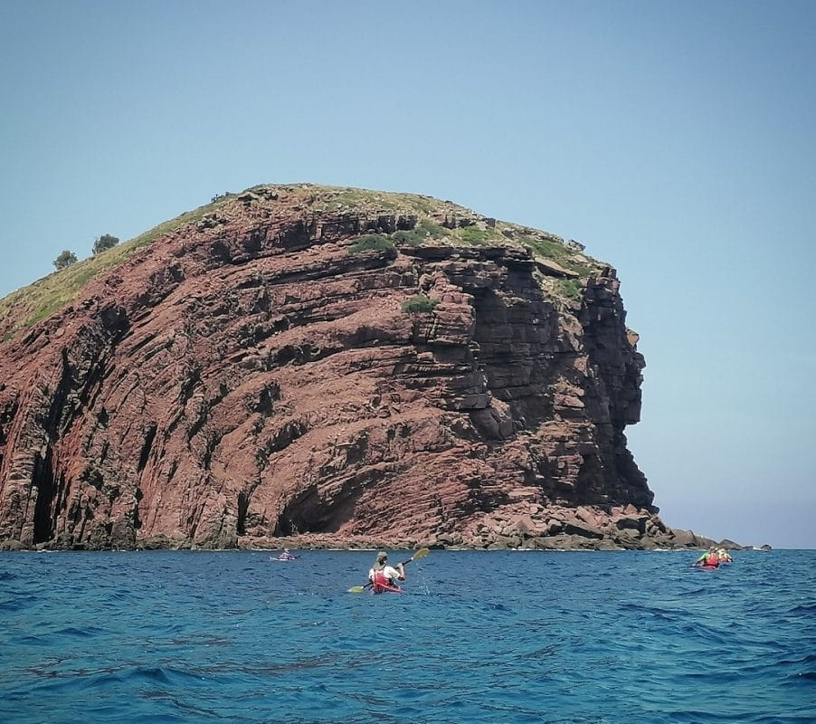 Tres personas en kayak reman por aguas azules frente a un gran acantilado rocoso de tonos rojizos en la costa de Menorca, bajo un cielo despejado.